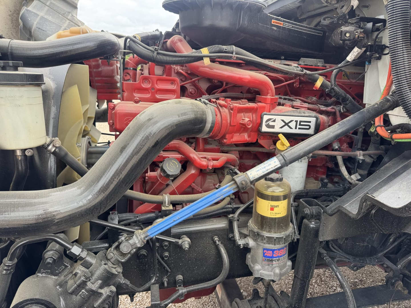 Truck engine bay with red engine labeled X15, large black intake hose, yellow coolant reservoir, and filter canister visible.