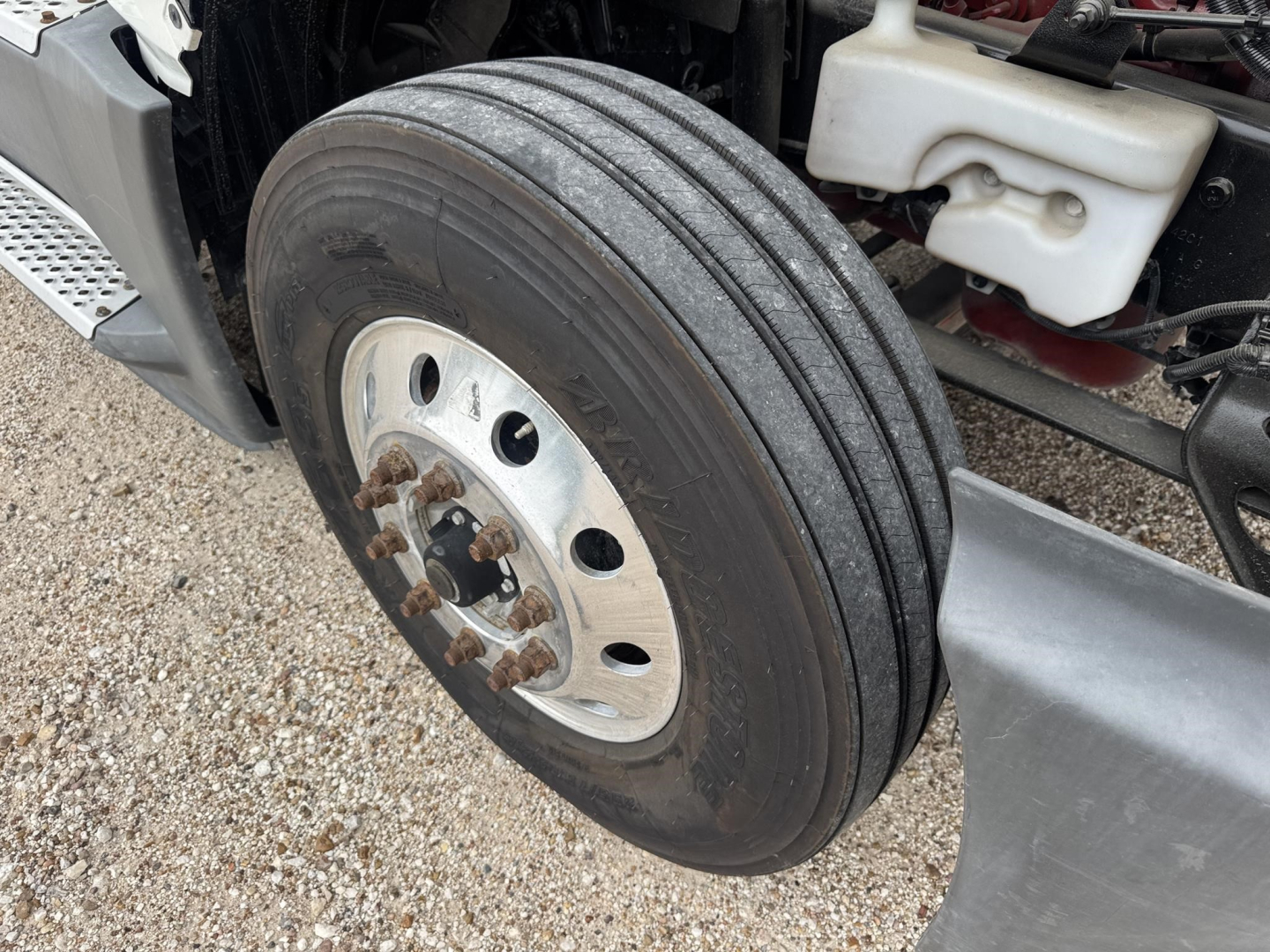 Truck front wheel close-up showing dual rear? no, single front tire with rim and lug nuts; parts include white plastic reservoir, metal steps, and chassis components visible.