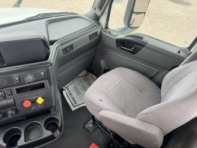 Truck cab interior showing steering wheel, driver seat, dashboard with controls, cup holders, and floor mats. Visible text on dashboard includes various control labels and a printed paper on the floor.