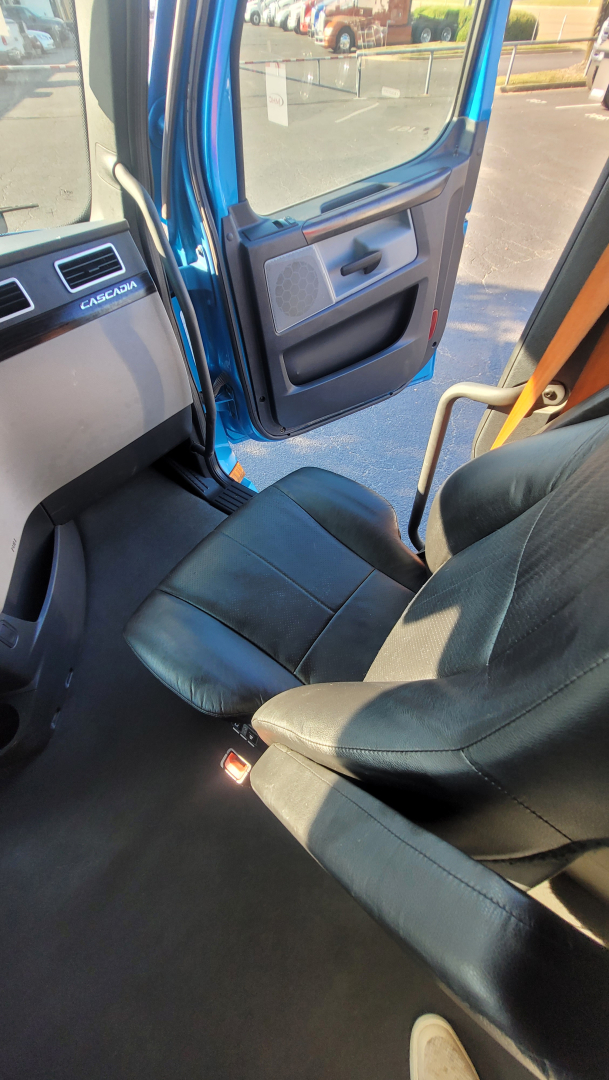 Cab interior of a Freightliner Cascadia 126 showing driver seat, door panel, and open door with blue exterior and floor mat.