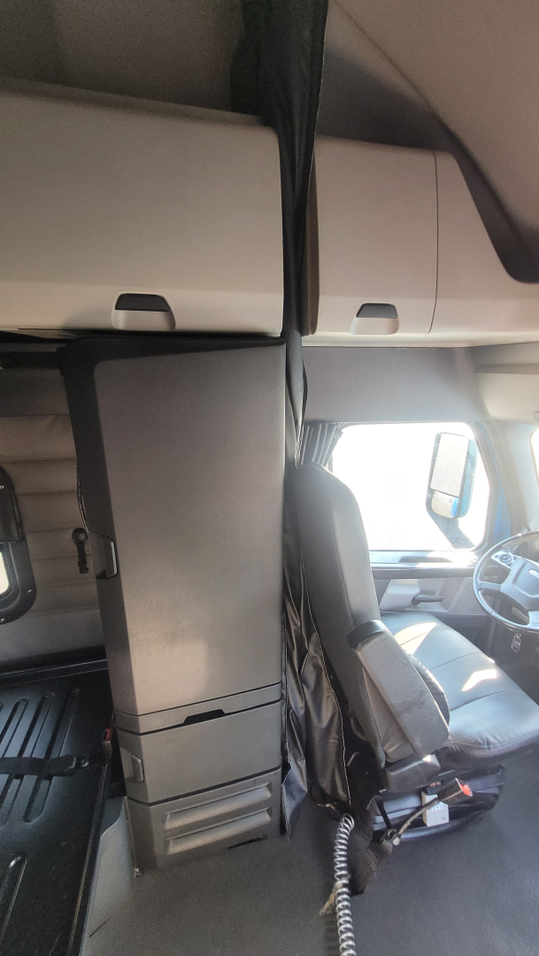 Cab interior of a Freightliner Cascadia 126 truck showing the driver's seat, center console, and overhead storage with a black partition.