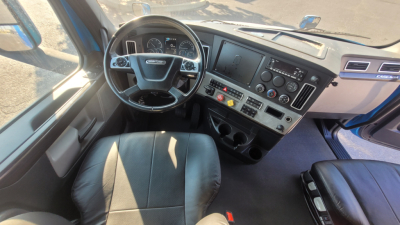 Truck cab interior showing steering wheel, dashboard, and multi-display panels in a Freightliner Cascadia with black seats.