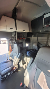 Cab interior of a 2022 Freightliner Cascadia 126 sleeper showing driver seat, passenger seat, overhead cabinets, and dashboard area with wiring conduit.