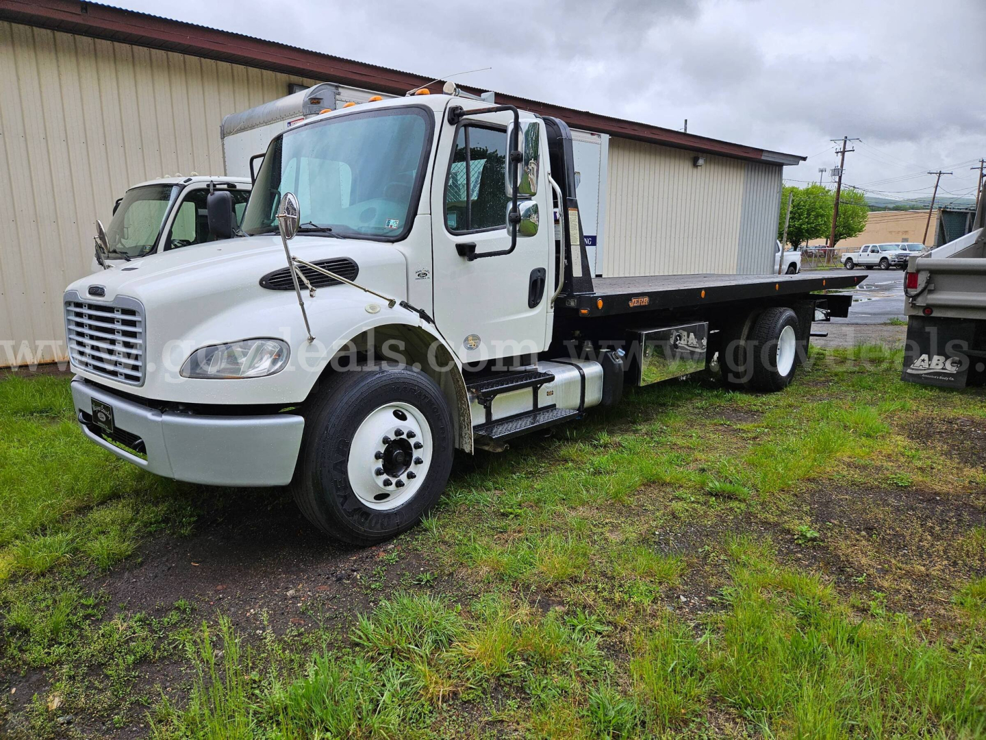 Flatbed tow truck front-left view with white cab and black flatbed, Freightliner M2 106 chassis, VIN tag area visible