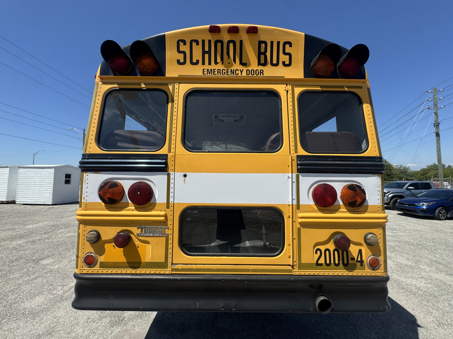 Rear view of a yellow school bus with black bumper, emergency lights, and '2000-4' on the lower right; 'SCHOOL BUS' sign at top; windows and taillights visible.