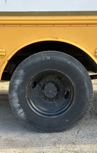 Partial side view of a yellow bus with a large black tire and wheel hub. No readable labels or serials visible on the wheel area.