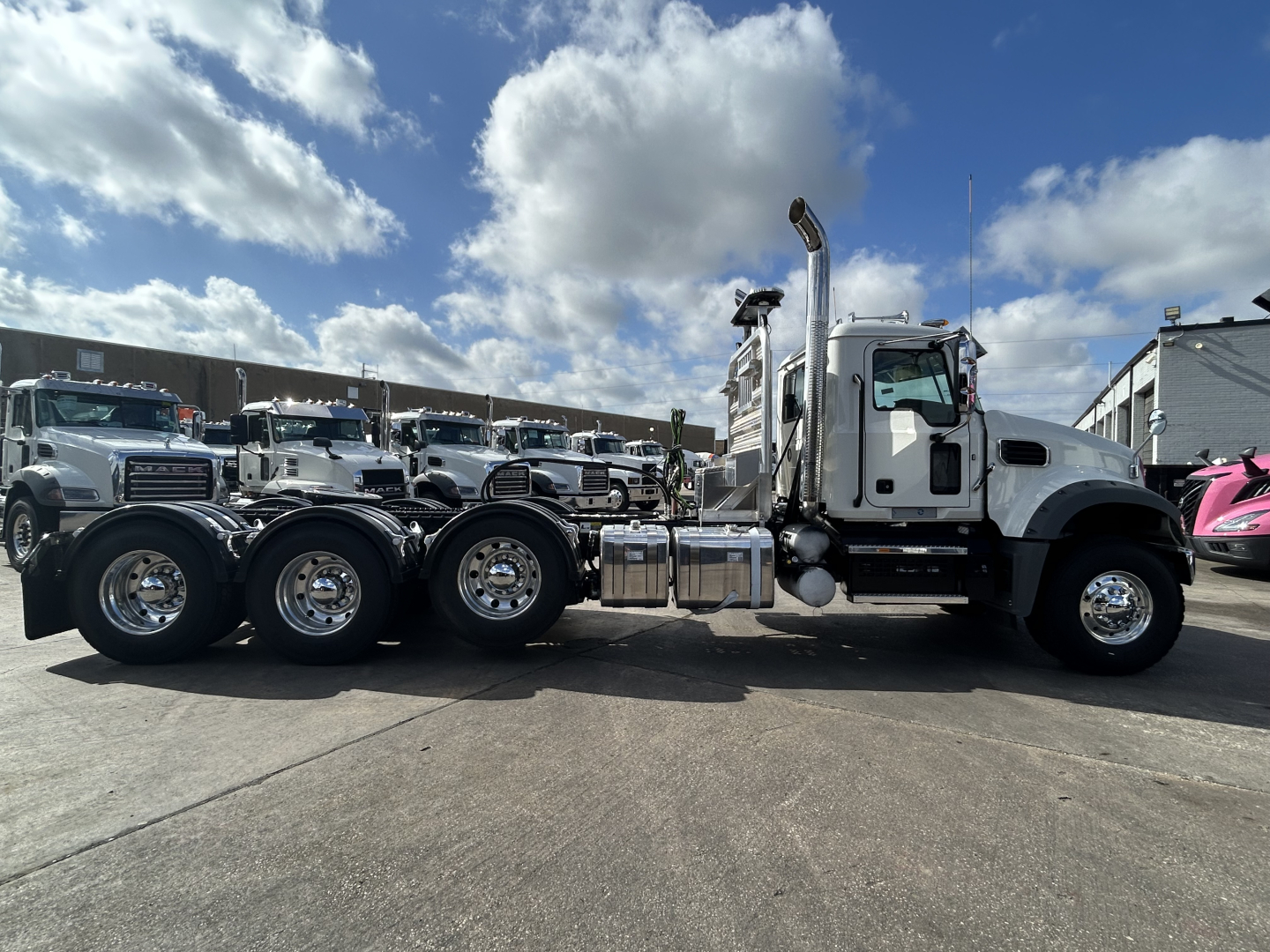 Mack GR84FT conventional daycab tractor showing chrome fuel tanks and rear axles, white cab, blue sky with clouds.