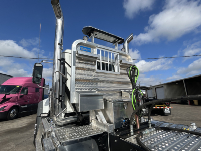 Front/side view of a 2027 Mack GR84FT conventional daycab tractor with polished aluminum tooling, diamond-plate steps, chrome exhaust stacks, and green and black hoses near the rear sleeper area.