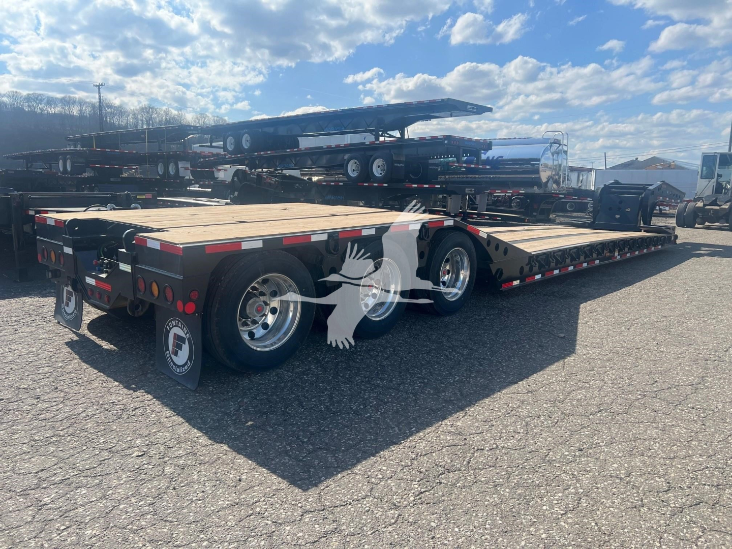 Fontaine paver trailer on a lowboy with tandem axles and wooden deck, viewed from rear-left, with other trailers in background.
