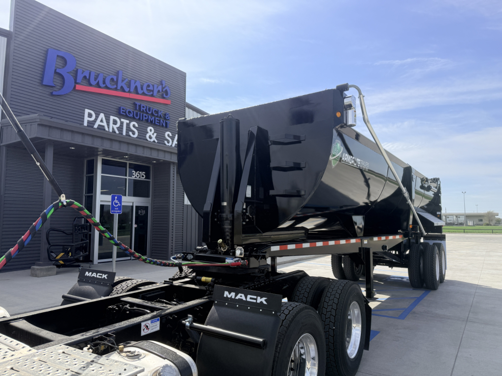 Dump trailer with 28/48 frame, Mack brand wheels, black glossy dump body, shown at a dealership lot, front tractor hitch in view.