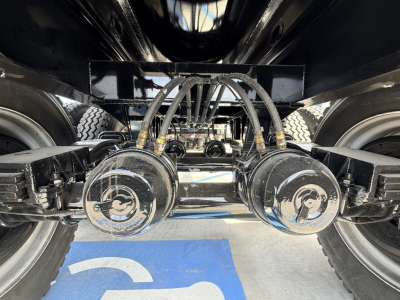 Trailer underside showing dual air brake chambers and hoses connected to axles, with wheels on either side and a blue floor marker.