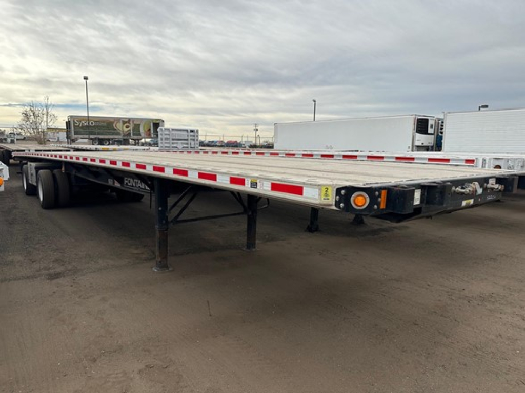 Flatbed trailer in a truck lot showing Fontaine branding on the frame and reflective tape along the sides. Dual rear axles and support legs visible.