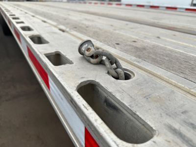 Flatbed trailer deck with a heavy-duty chain anchor and recessed tie-down slots showing exterior aluminum surface.