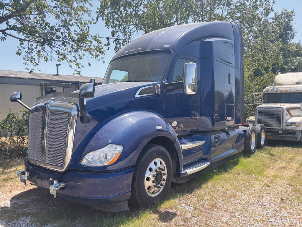 Blue 2019 Kenworth T-680 conventional sleeper tractor, front three-quarter view, with chrome grille, dual rear axles, and a white other truck in background.
