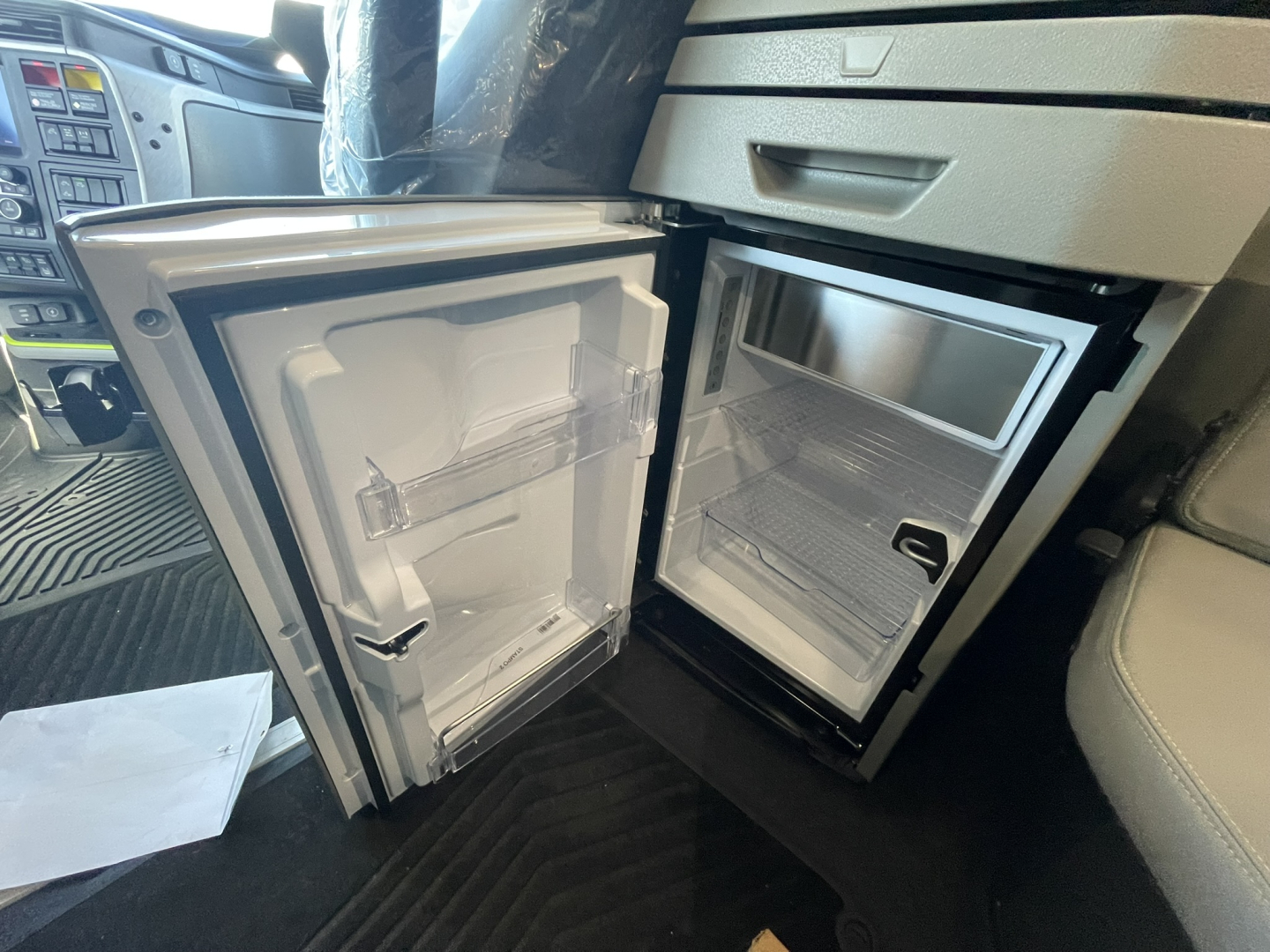Interior of a Volvo VNL truck cab showing an open compact refrigerator/freezer unit with empty shelves and a small freezer drawer. Dashboard and steering area visible in background.