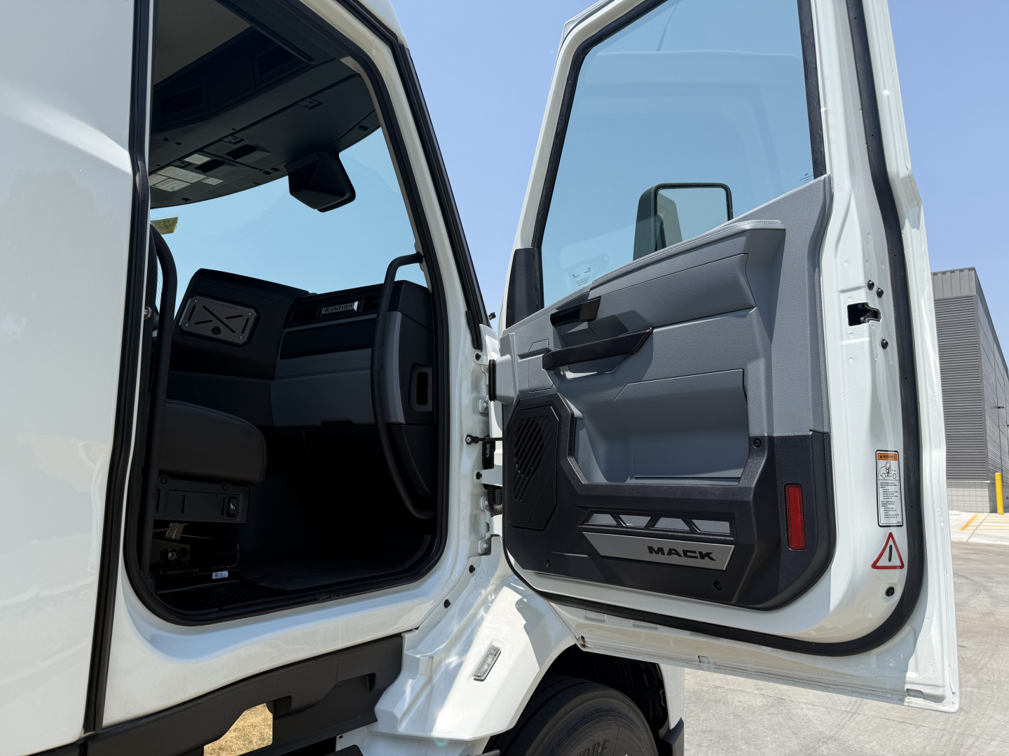 Truck cab interior view with open doors of a 2027 Mack AN64T, showing driver and passenger door panels, step area, and steering wheel visible inside.