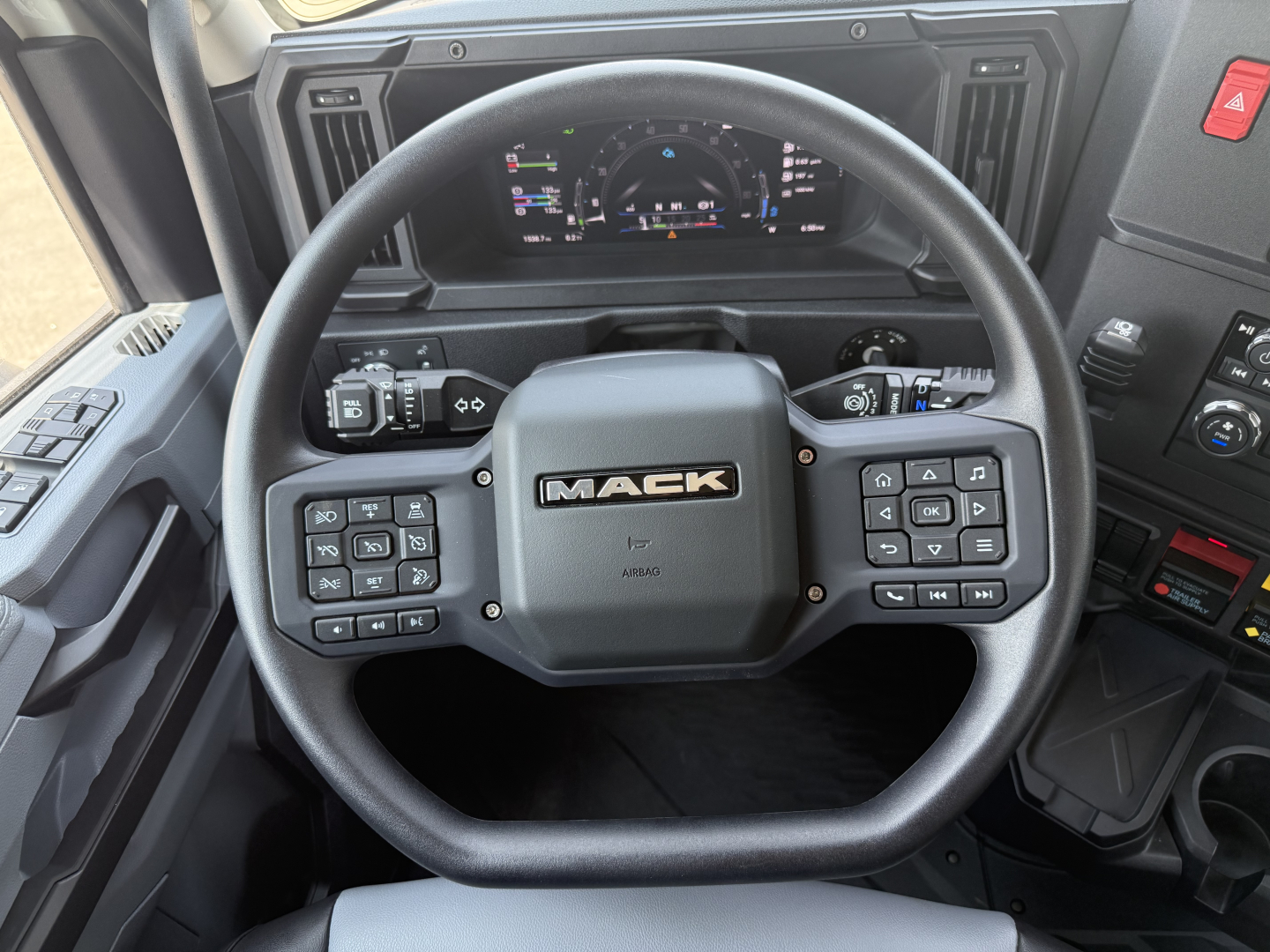 Mack AN64T cabin interior showing steering wheel with Mack logo and control buttons, instrument cluster visible behind wheel, dashboard switches on both sides.