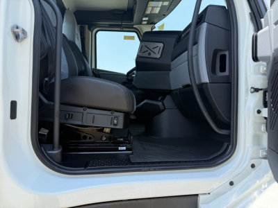 Cab interior of a new Mack AN64T daycab truck showing driver seat, passenger seat, and entry doorway; visible controls and flooring, light-colored exterior frame.