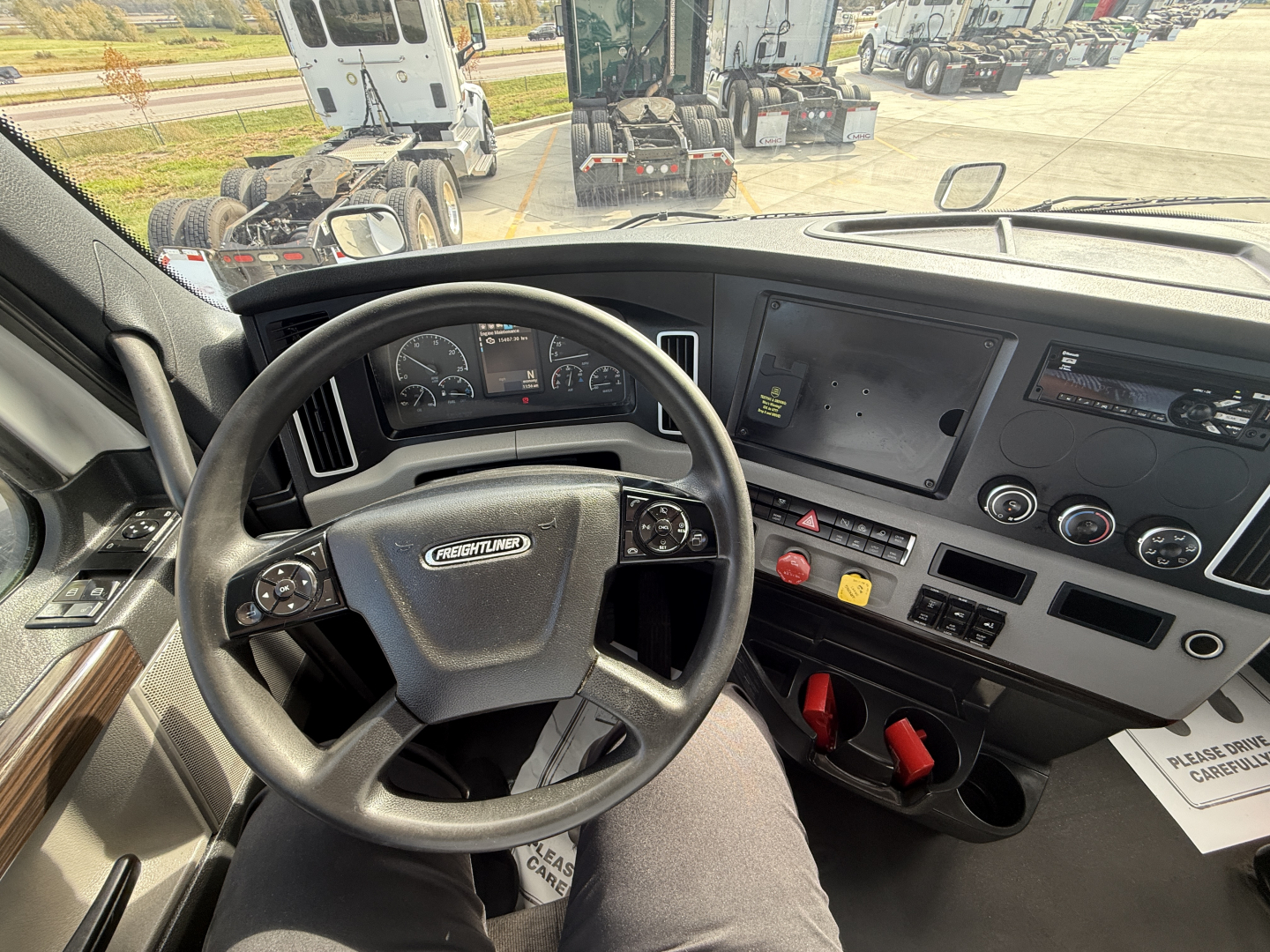 Cab interior of a Freightliner truck showing steering wheel, instrument cluster, central display, and control panel. Visible are red tractor controls and cupholders.
