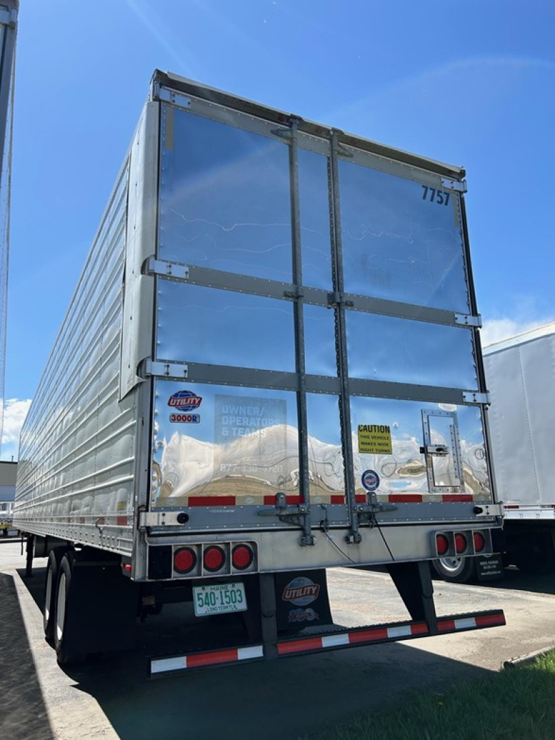 Trailer rear view showing aluminum-sided Utility 3000R Reefer trailer with dual rear doors, taillights, and liability stickers. Visible numbers 7757 on door, rear bumper, and reflective tape.