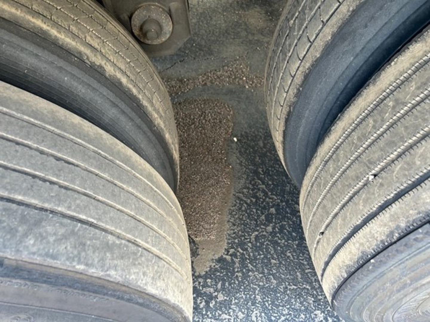 Rear dual-axle trailer tires with a view of the muddy ground between the wheels and a portion of the trailer frame.