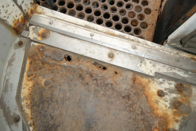 Rusted metal floor and perforated step of a truck cab; visible rust and wear around metal edge, with perforated grate in back.