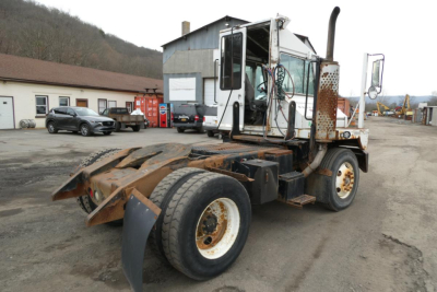 Tractor truck chassis in yard showing cab and dual rear wheels with rusty metal hitch components and exhaust stack.
