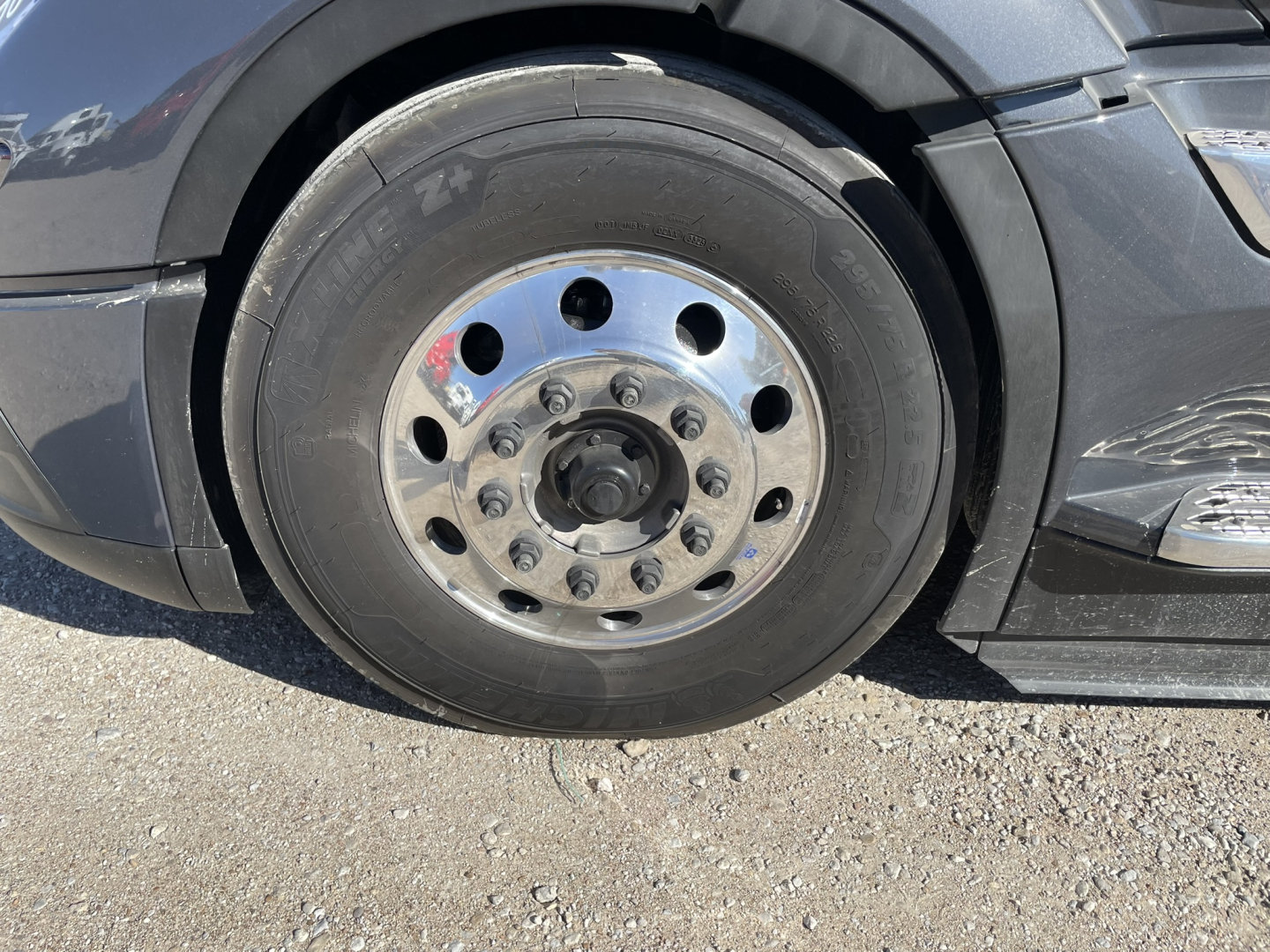 Volvo heavy truck front wheel showing steel rim with multiple lug nuts and black tire. No visible text on tire; wheel well and side panel visible.