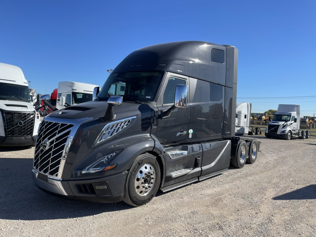 Volvo VNL64T860 tractor unit in a dealership lot, chrome grille, black cab, sleeper, three axles, front wheel, side mirrors, various trucks in background.