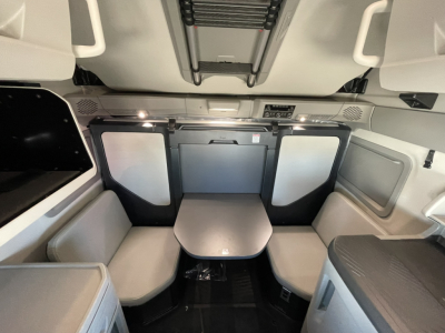 Interior of a Volvo VNL64T860 cabin showing a small table between two bench seats with light gray and black trim, overhead controls, and storage panels.