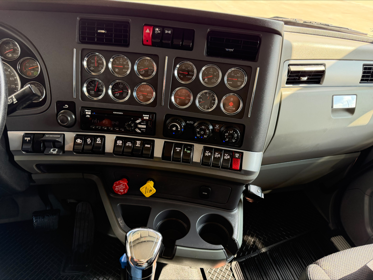 Dashboard and instrument panel of a Kenworth T880 cab showing multiple gauges, switches, HVAC controls, cup holders, and gear shifter; center console with various indicator labels and knobs.