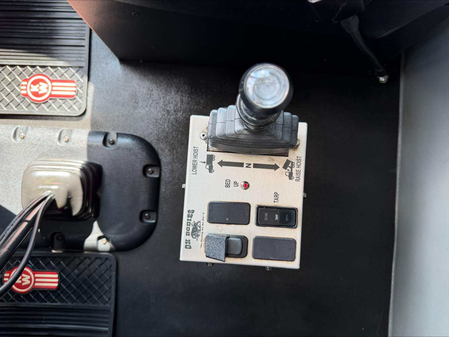 Kenworth T880 dash area showing forklift-style shift lever and control panel with multiple buttons and labels, near pedals and textured panels.
