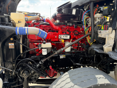 Kenworth T880 engine bay showing bright red X15 engine with blue radiator hose, brake reservoir, wiring harnesses, and surrounding components.