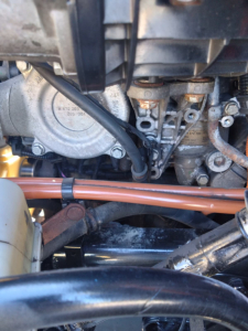 Truck engine bay view with metal engine components, hoses, and a circular housing. Visible small engraved numbers on metal surface. Close-up of mechanical fittings and wiring.