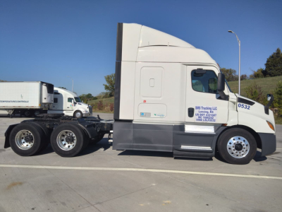 Freightliner Cascadia truck side view showing sleeper cab with white/gray color, wheels, and hitch; visible text on sleeper door and side panel includes company branding and contact details.