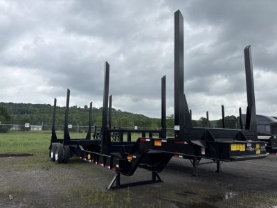 Log trailer with tall vertical stakes, black steel frame, tandem axles, and gooseneck hitch showing at an outdoor yard.