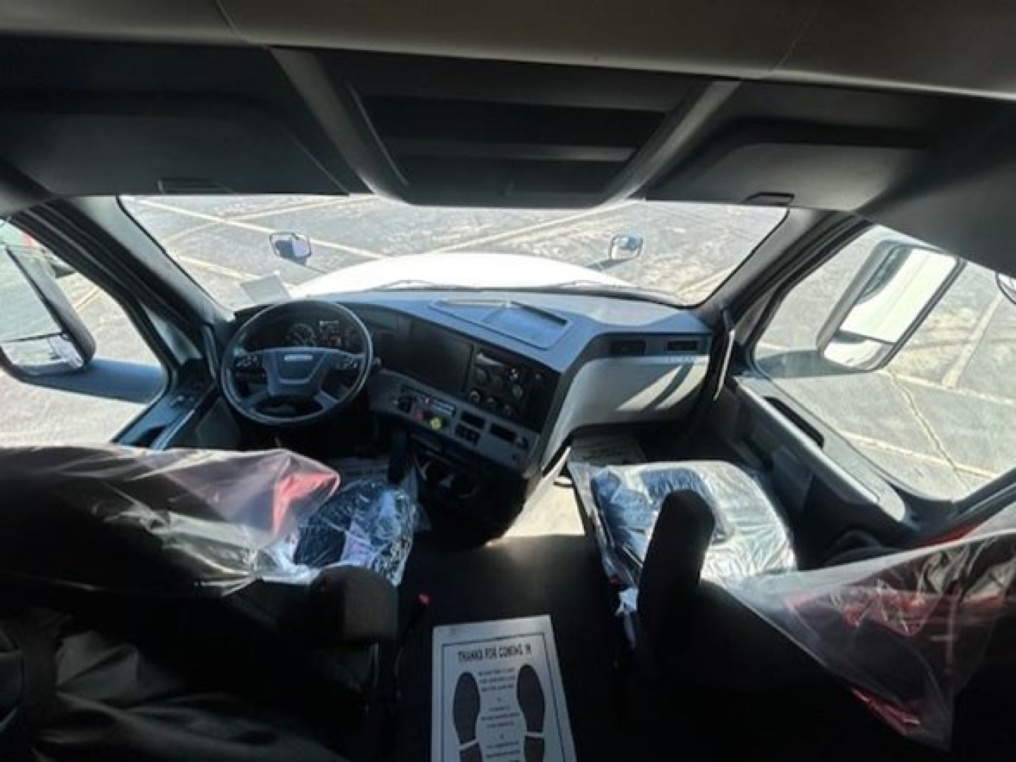 Truck cab interior of a Freightliner Cascadia with plastic seat covers, dashboard cluster, steering wheel, and front seats visible.
