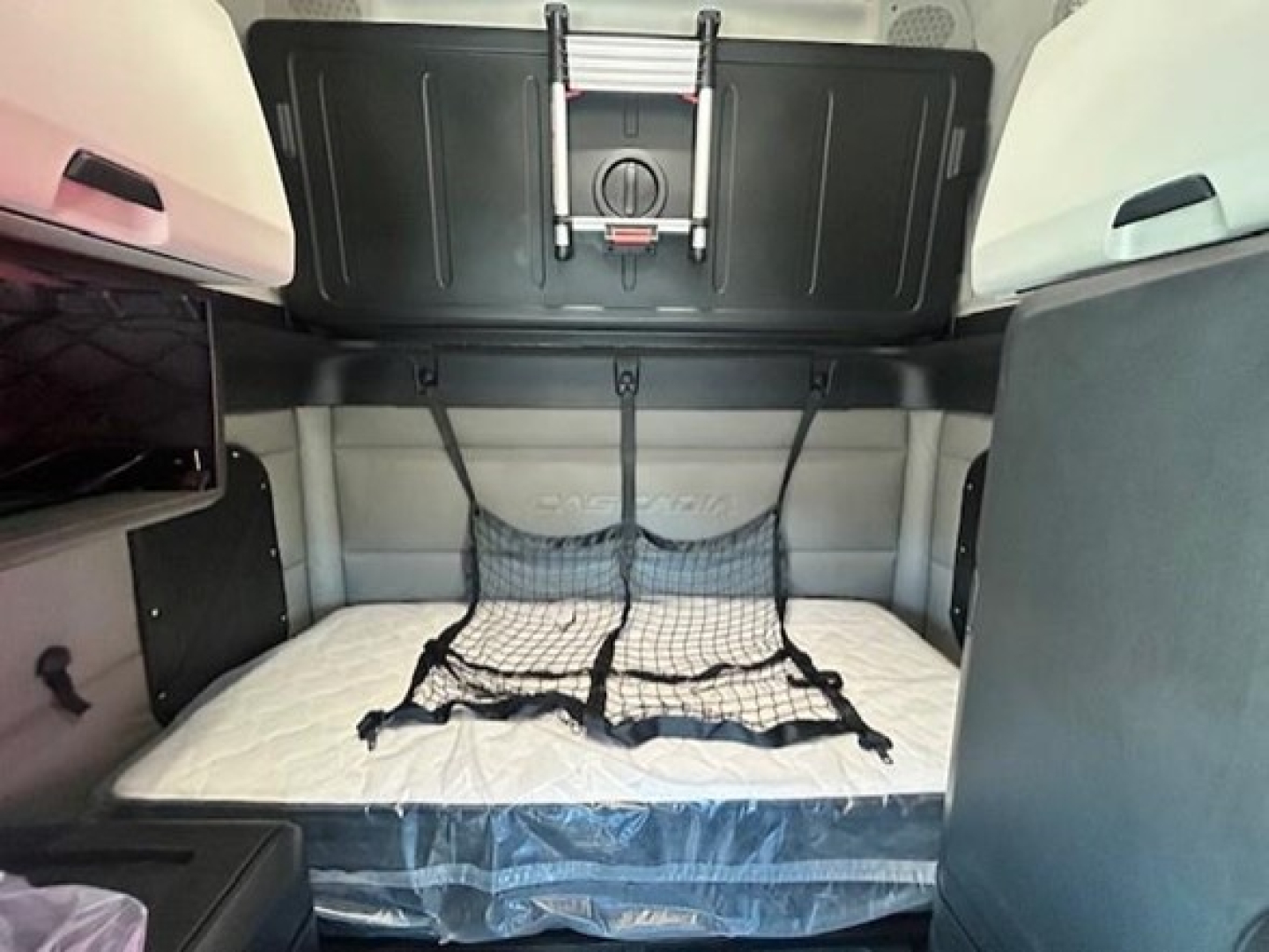 Interior sleeper cab of a Freightliner Cascadia showing a raised bunk area with a netted storage and overhead cabinet.