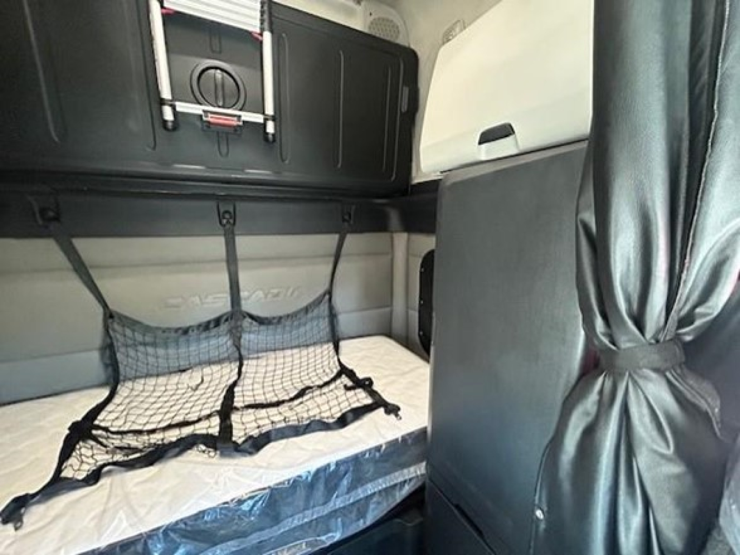 Freightliner Cascadia sleeper cab interior with bunk area and storage netting, visible mattress, overhead compartments, and curtain.
