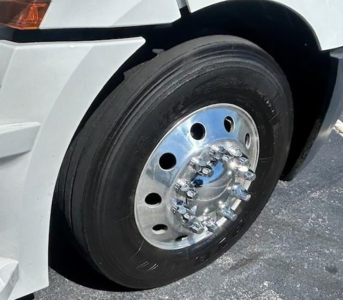 Front wheel of a Freightliner Cascadia 126 showing a steel hub with lug nuts and black tire, part of a white truck fender.