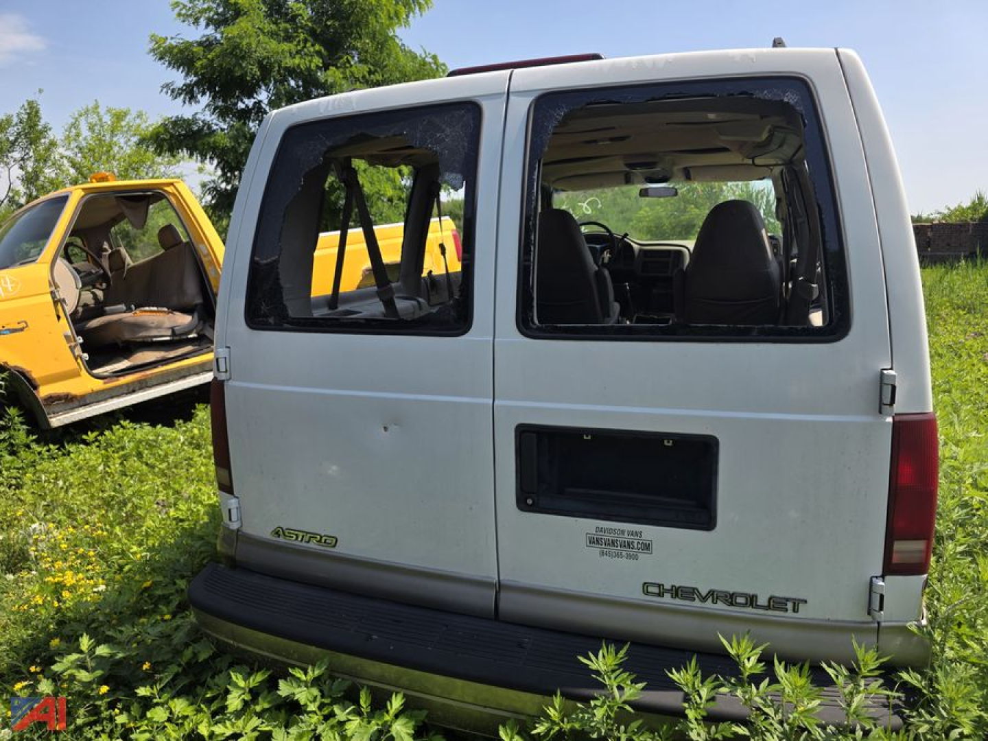 Chevrolet Astro van rear view showing two doors, missing rear window panes, and fitness of a white cargo van with Chevrolet emblem. Surrounding vegetation and a yellow vehicle in background.