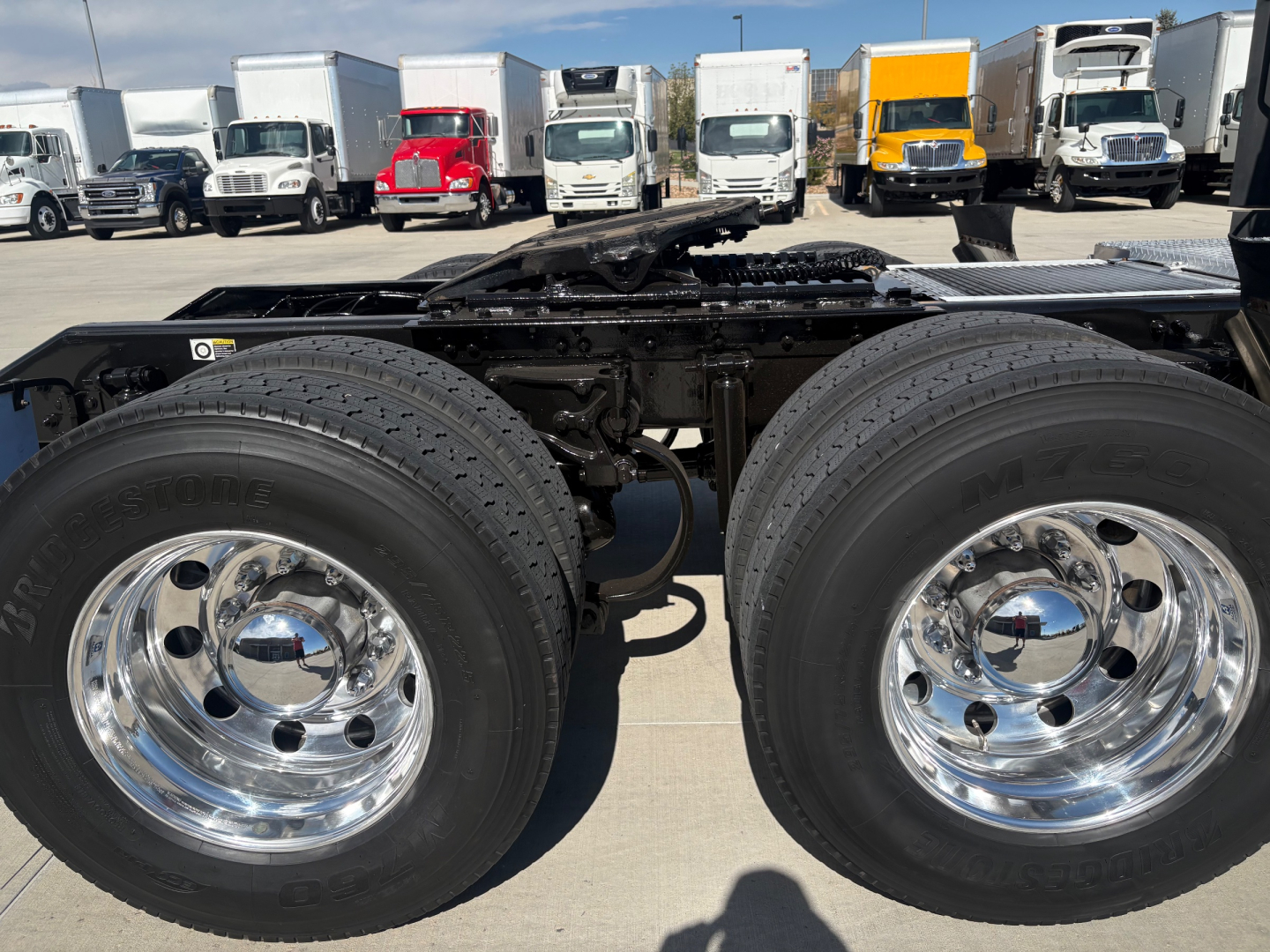 Kenworth T680 chassis with dual rear dual tires, chrome wheels, and part of cab visible; background shows a row of trucks in a dealership lot.