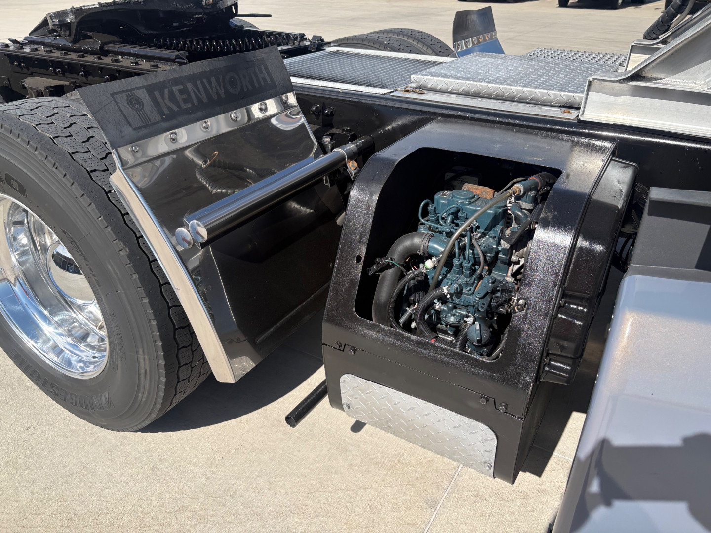 Truck chassis side area showing a diesel engine under a hinged cover, Kenworth branding visible on a panel, metal steps, and rear wheel with chrome rim.