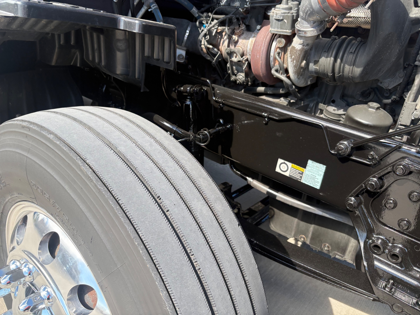 Kenworth T680 chassis with front dual drive? view showing front tire and engine bay components. Visible components include suspension crossmember, hydraulic lines, and engine bay hardware.