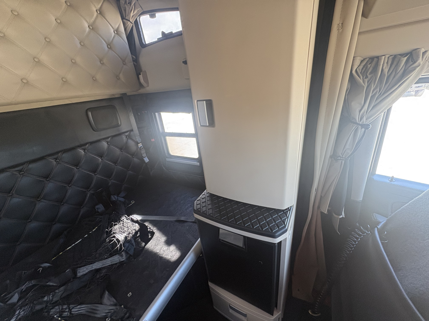 Interior sleeper area of a 2022 Kenworth T680 truck showing a vertical partition, small cabinet, and quilted upholstery on the wall. Window and curtain visible; sunlight entering.