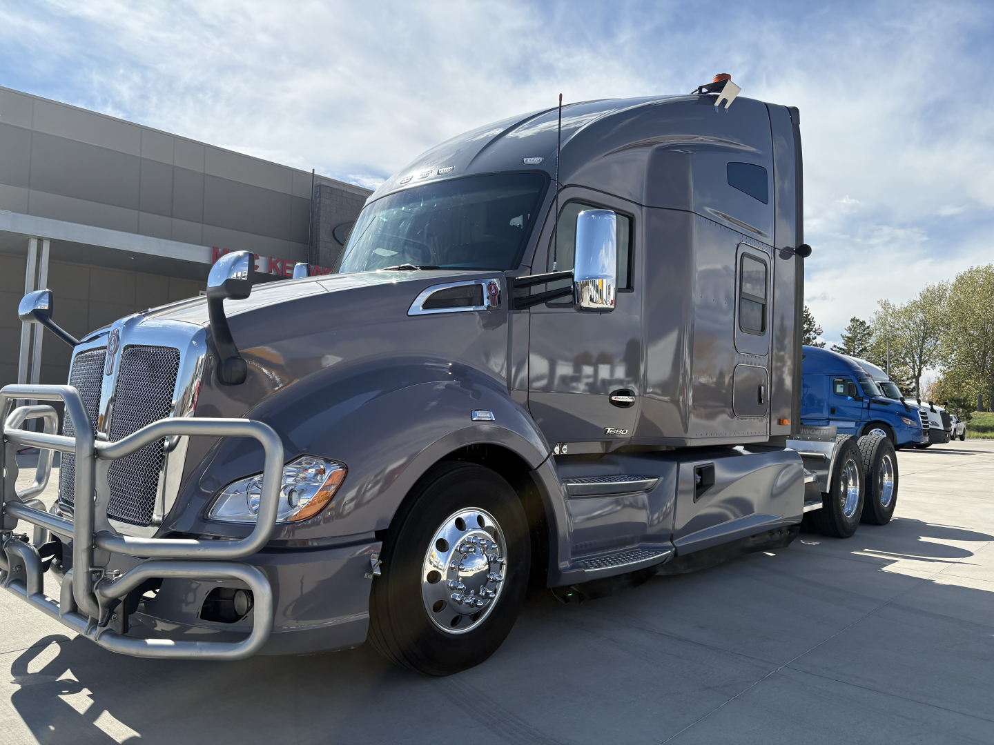 Kenworth T680 tractor cab in a parking lot, front grille with bumper guard and side mirrors, chrome wheels; no legible labels visible on the truck from this angle.