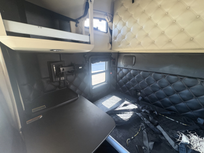 Kenworth T680 sleeper cab interior view showing black quilted door panels, overhead shelf, small window, and workstation with wiring harnesses and a black countertop.