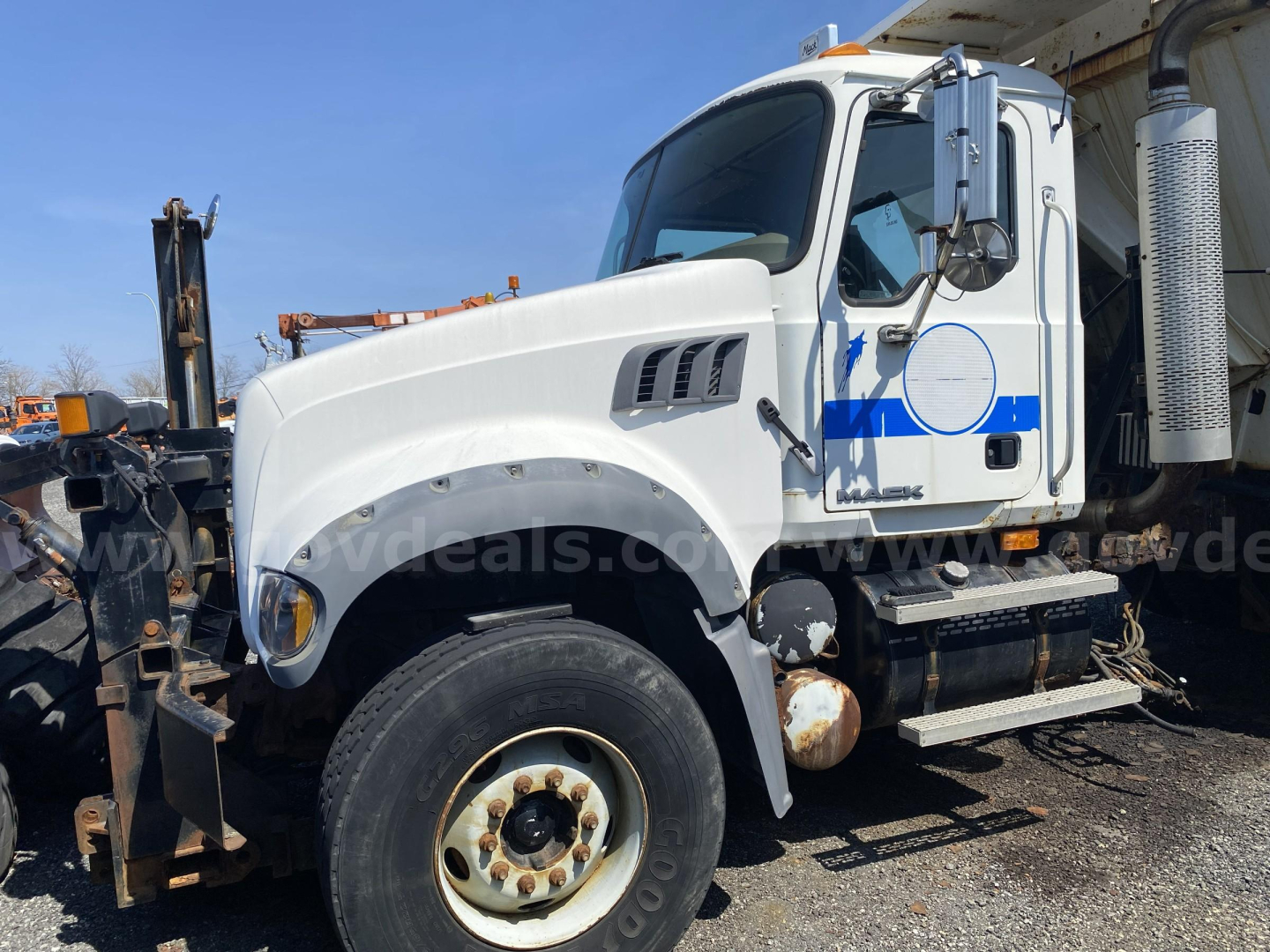 Truck cab side view showing Mack CTP713 dump truck with white paint and blue stripe, front wheel, exhaust stack, and equipment attachments.