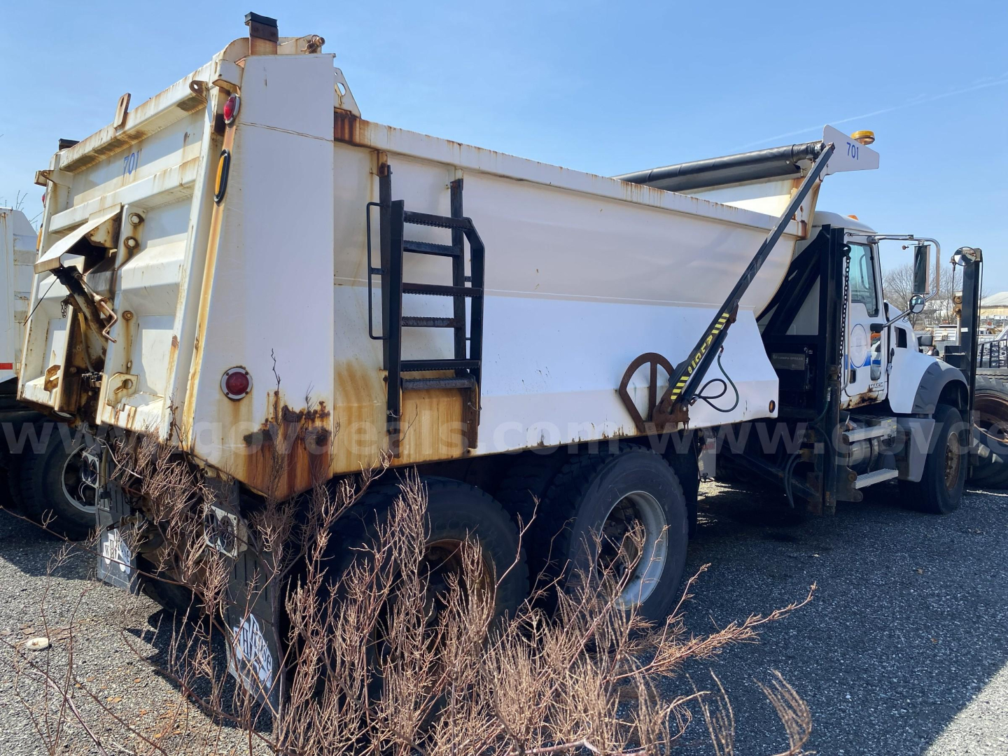 Dump truck bed showing hydraulic system and rear dual tires with rusted edge and ladder on side.