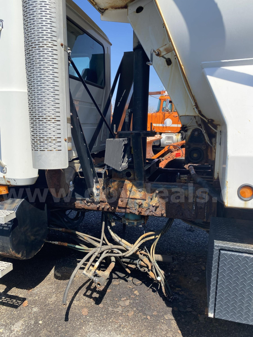 Truck engine bay area with hydraulic hoses and dump bed linkage visible between cab and dump body. Mack CTP713, side view, metal components, hoses, and brackets.